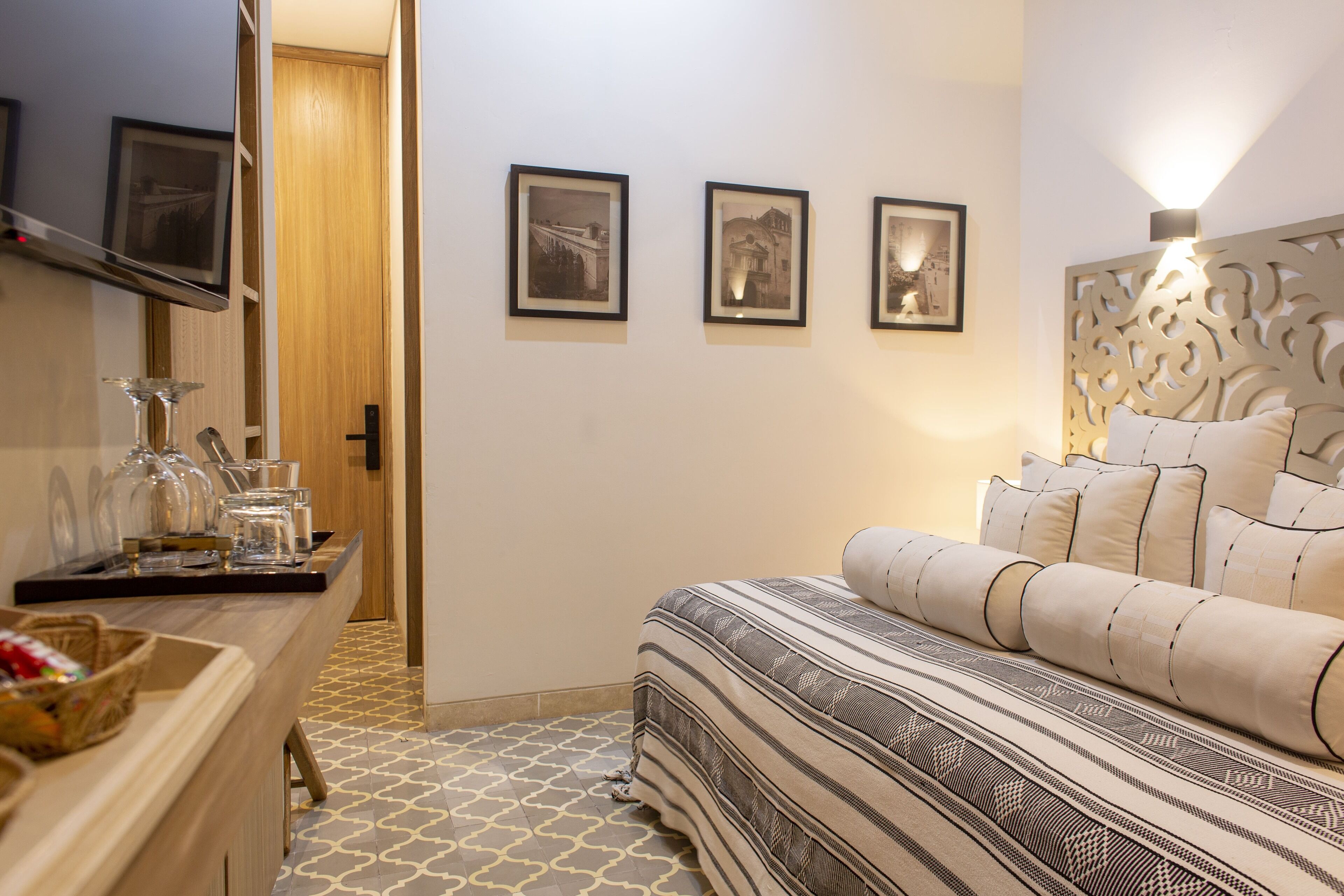 A bedroom with a bed, a bedside table with glasses and a basket, and three framed pictures on the wall.