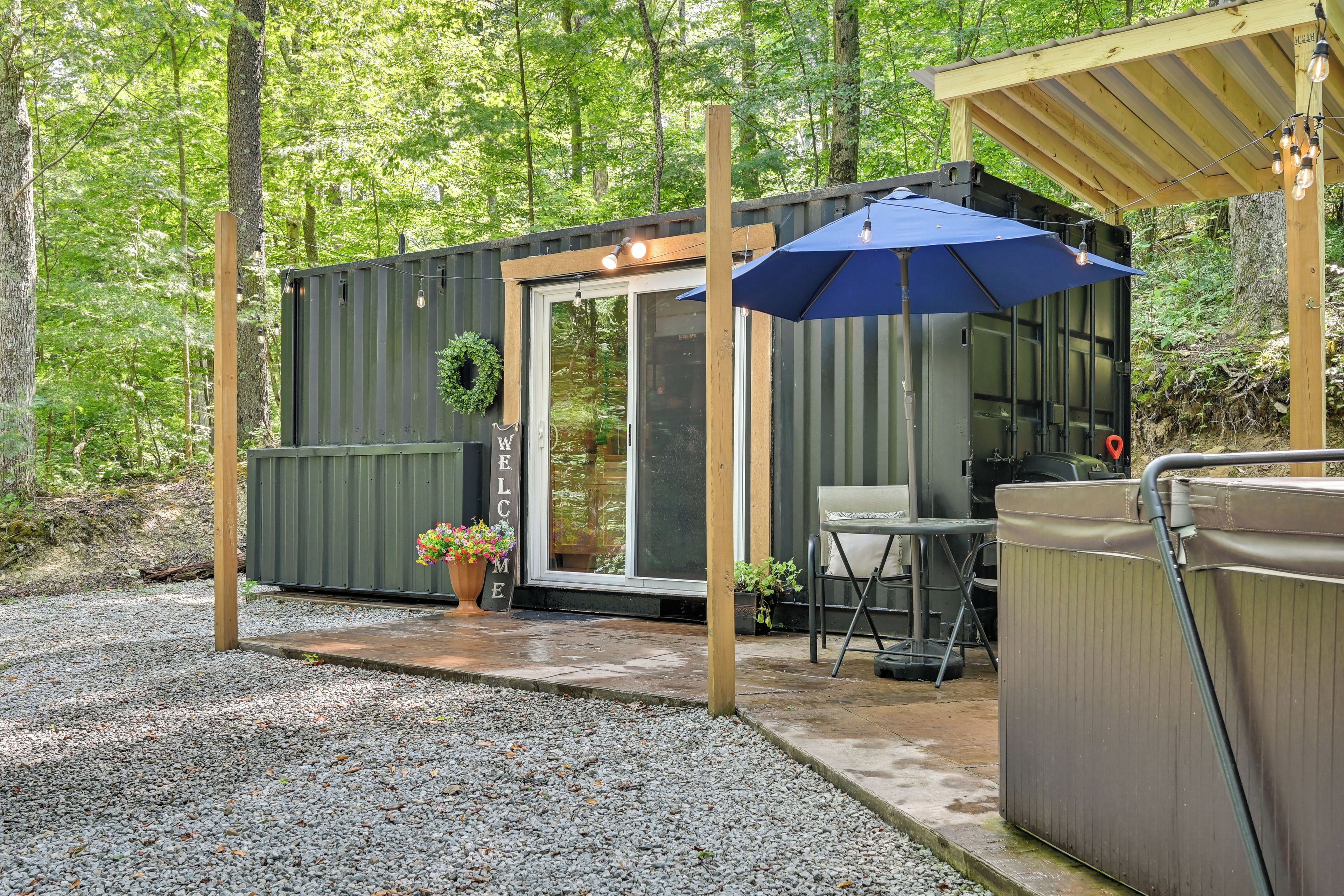 A wooden deck with a hot tub, a table, and chairs, surrounded by a gravel area and a metal structure with a blue umbrella.