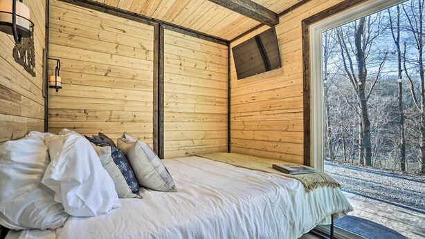 Un dormitorio de cabaña de madera con cama, almohadas, una ventana y vistas a árboles.