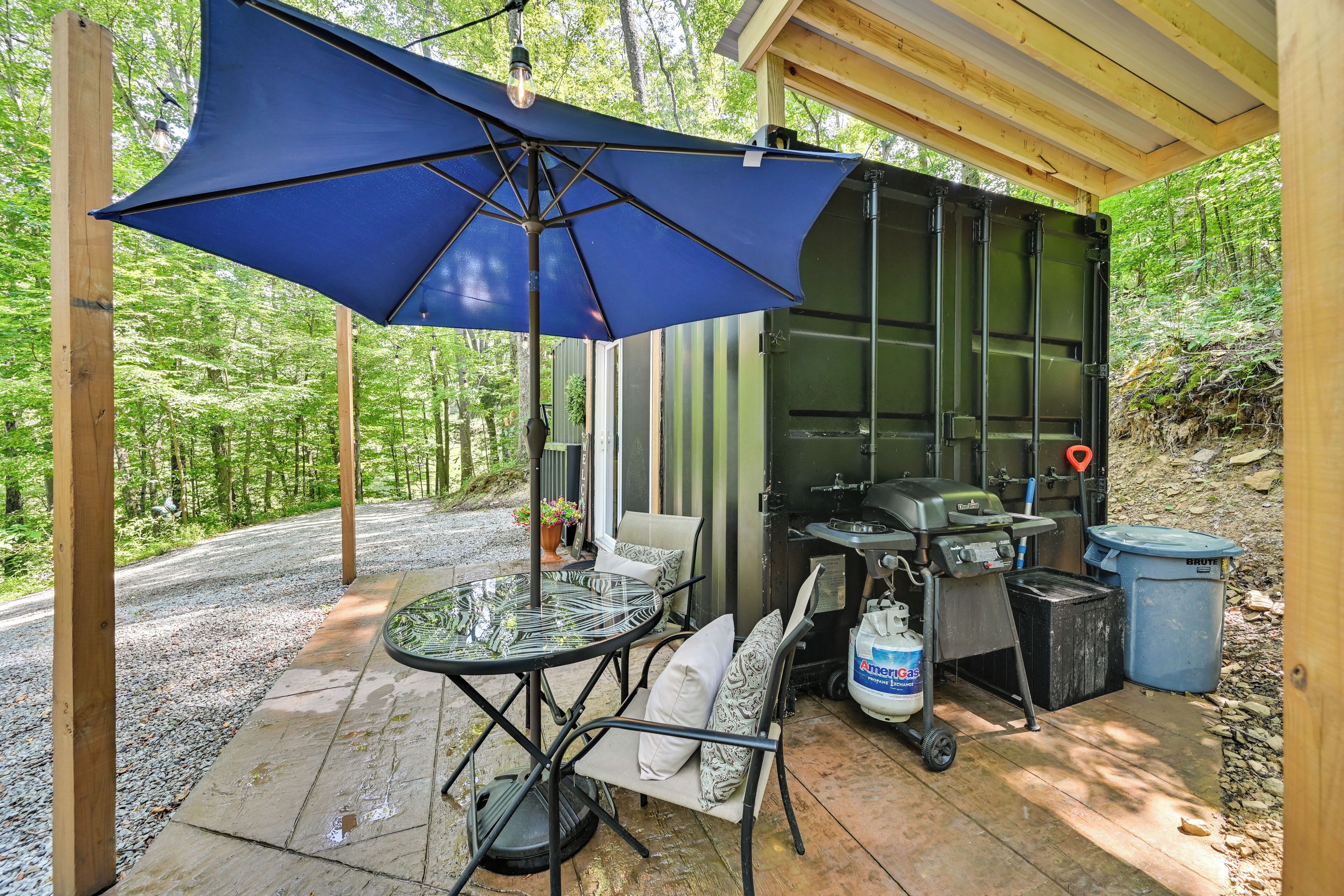 An outdoor patio with a table and chairs, a blue umbrella, a grill, and a trash can.