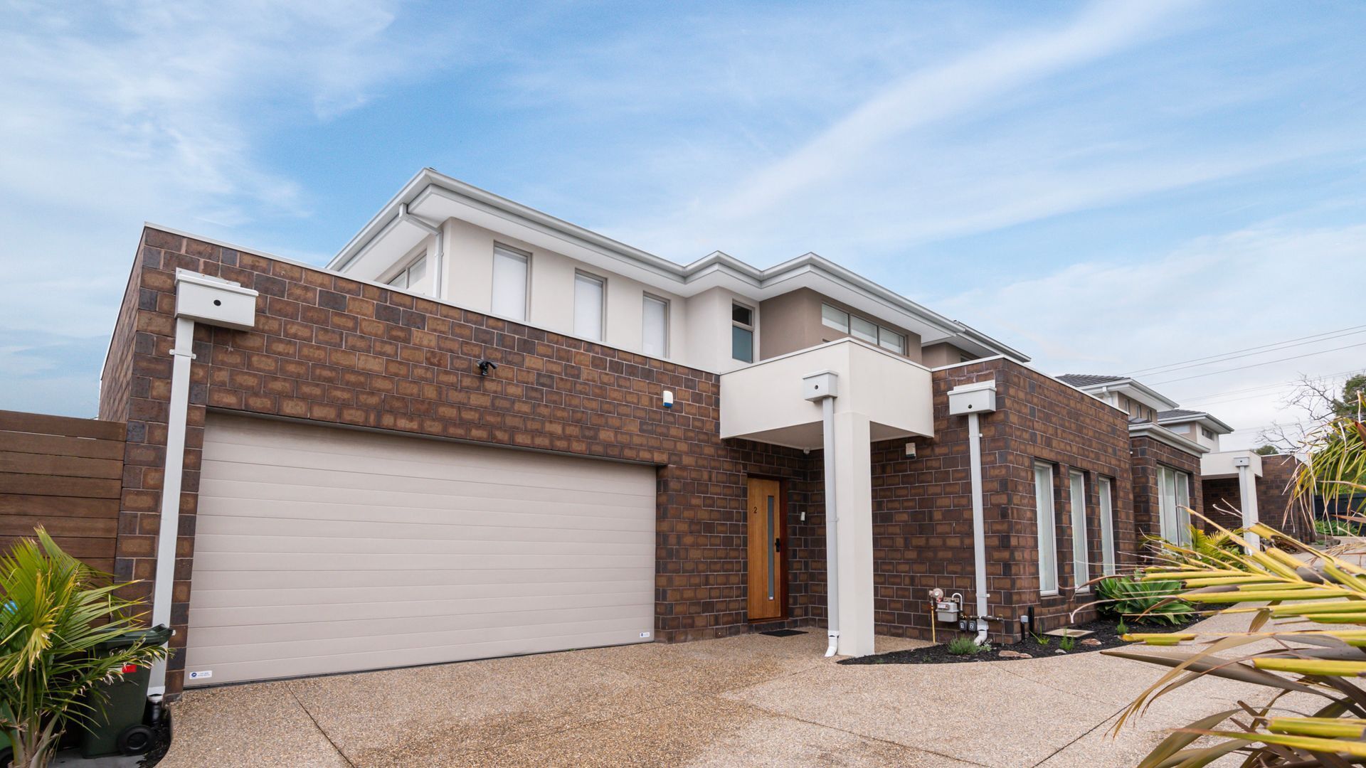 A modern two-story house with a garage, brick facade, and a small front yard with plants.