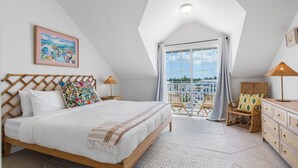 A bedroom with a wooden bed, a colorful pillow, a nightstand, a lamp, a wicker chair, a dresser, and a balcony with a view. - Convenient Stay in the Heart of Sandyport Olde Towne (Nassau)