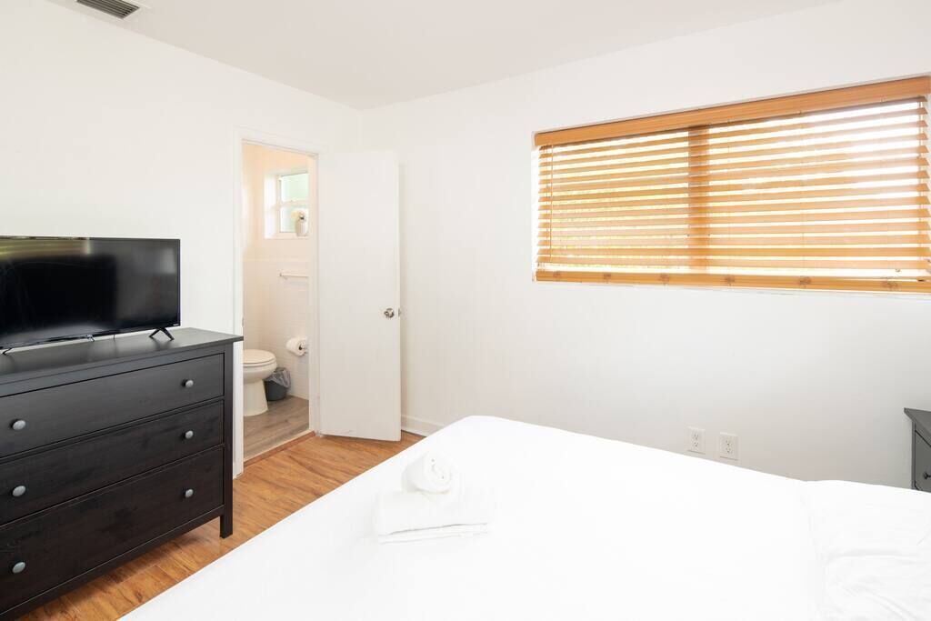 A bedroom with a bed, a dresser, a television, a window with blinds, and a door leading to a bathroom.