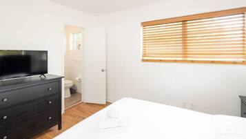 A bedroom with a bed, a dresser, a television, a window with blinds, and a door leading to a bathroom.