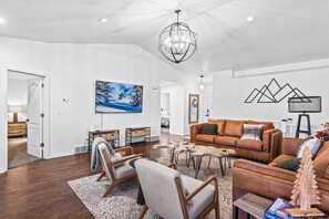 A spacious living room with a brown leather sofa, a coffee table, and a chandelier. There is a painting on the wall and a wooden sculpture on the coffee table. - Casa Cliffside: Animal Friendly, Mountain Getaway w/ Private Balcony (Logan)