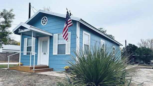 Una piccola casa blu con una porta bianca, una bandiera americana e una grande pianta di agave sullo sfondo.