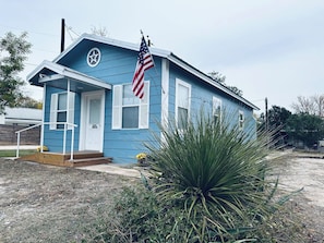 A small blue house with a white door, an American flag, and a large agave plant in the foreground.
