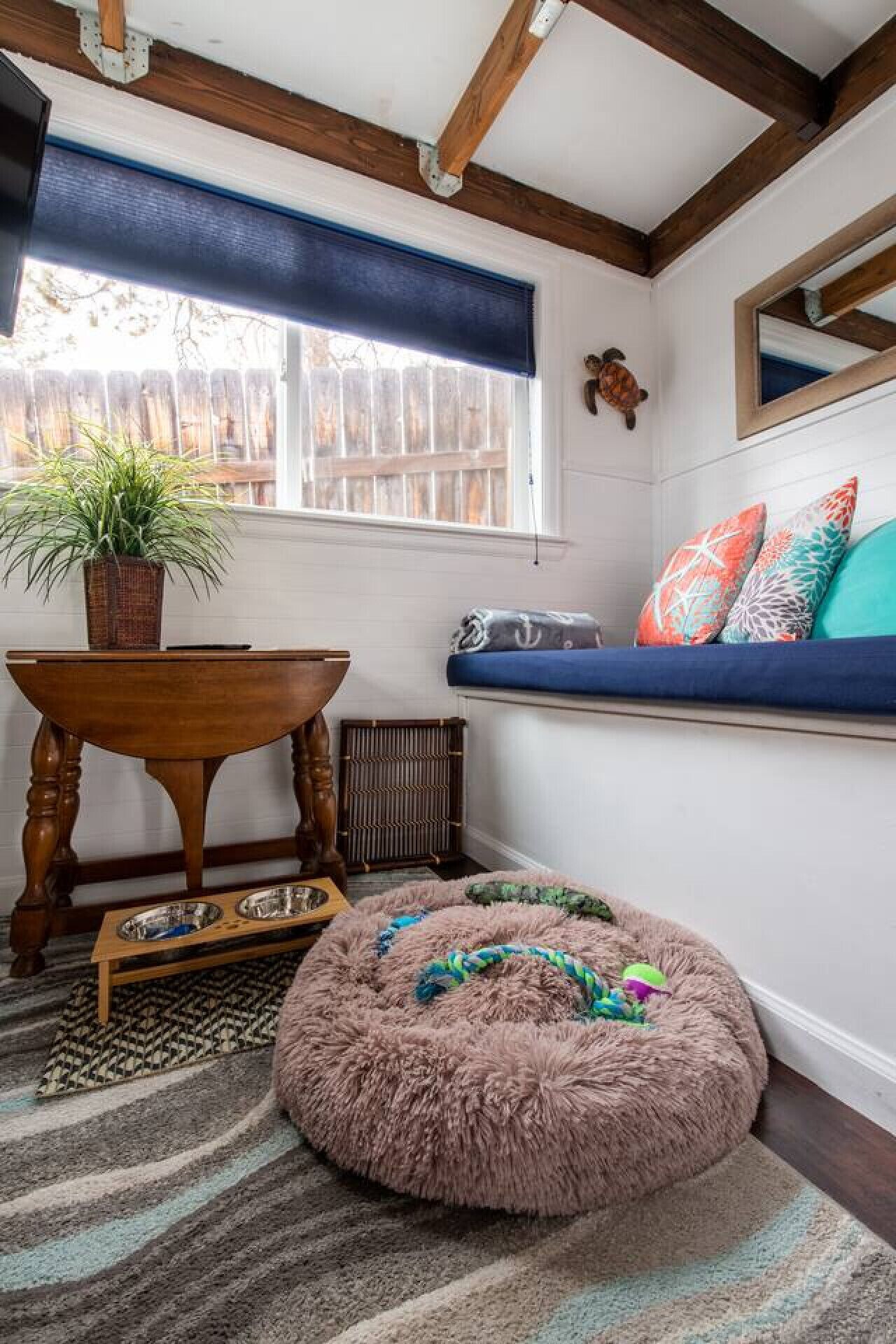 A cozy room with a wooden table, a fluffy pet bed, a window with a view, and a built-in bench with colorful pillows.