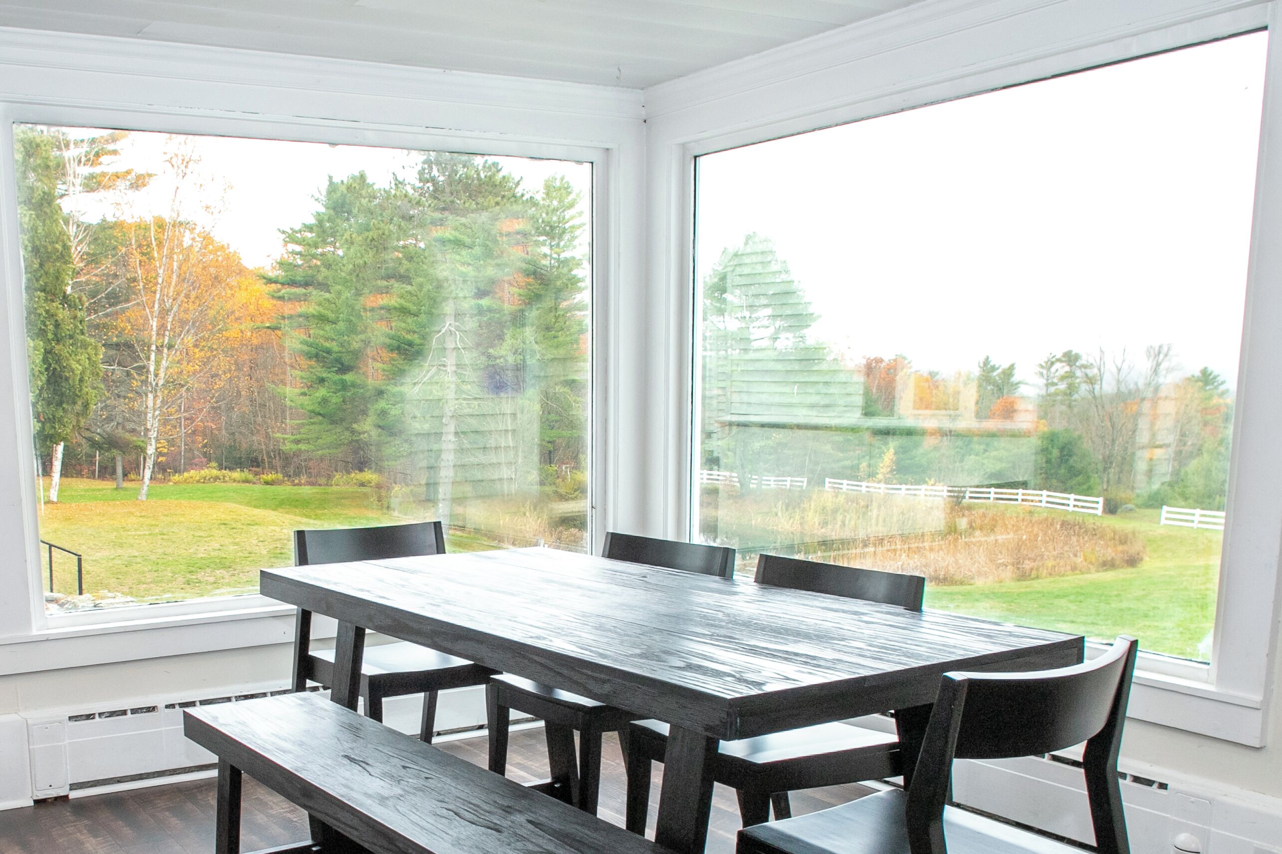Un espace repas avec une grande table et des chaises, de grandes fenêtres donnant sur une cour verdoyante et des arbres.