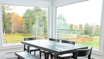 Un espace repas avec une grande table et des chaises, de grandes fenêtres donnant sur une cour verdoyante et des arbres.