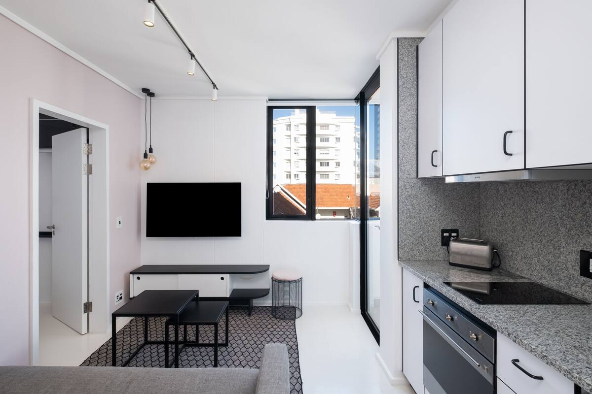 A modern kitchen with a flat-screen TV, a dining area with a bench, and a view of a building through a large window.
