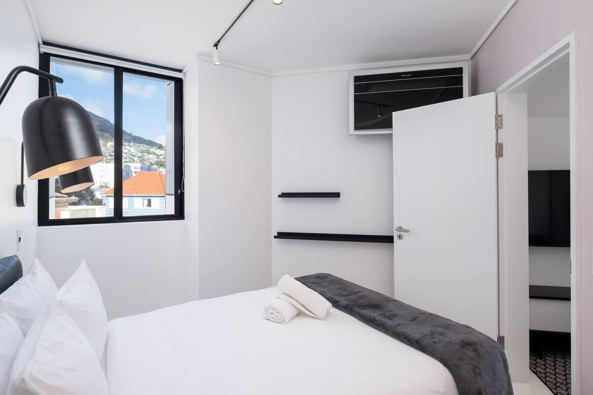 A modern hotel room with a bed, a window with a view of buildings and mountains, a black lamp, and a white door.