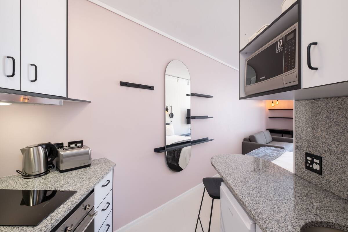 A modern kitchen with a microwave, toaster, and kettle. There is a mirror on the wall and a countertop with a sink.