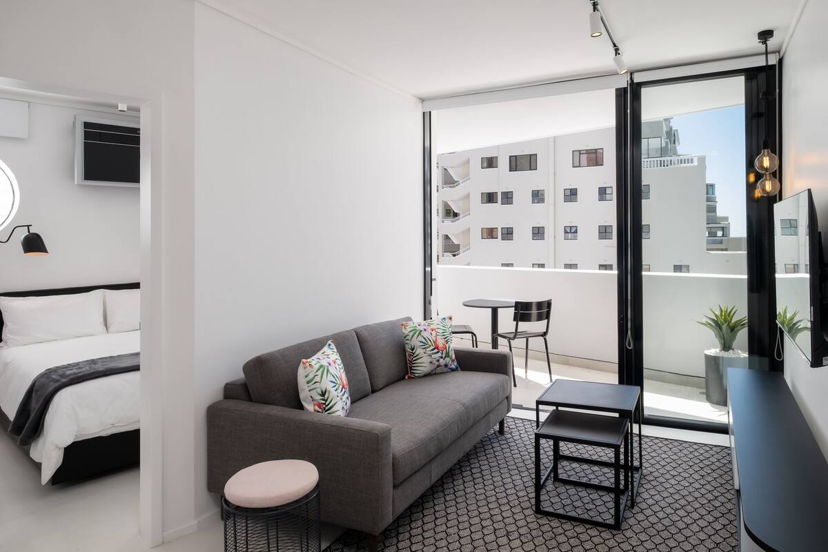 A modern hotel room with a grey sofa, a small round table, a bed with white bedding, and a large window offering a view of the cityscape.