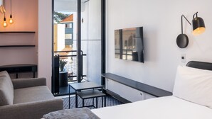 Une chambre d’hôtel moderne équipée d’un lit, d’un canapé, d’une petite table, d’une télévision et d’un balcon offrant une vue sur les bâtiments.