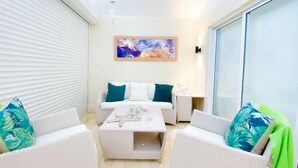 Eine sonnendurchflutete Terrasse mit einer weißen Couch, türkisfarbenen Kissen, einem Couchtisch, Korbstühlen und einer Glastür, die zu einem Balkon führt.