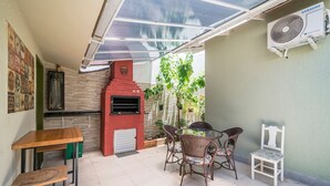 A patio area with a red brick oven, wicker chairs, a table, and a Samsung air conditioning unit. - 918 - Casa grande 3 Quartos 8 pessoas (Bombinhas)