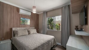 A bedroom with a bed, an air conditioning unit, a painting, a window with curtains, and a television on a stand. - 918 - Casa grande 3 Quartos 8 pessoas (Bombinhas)
