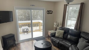 A living room with a leather sofa, a black ottoman, a wooden coffee table, and a TV on the wall. There is a sliding glass door leading to a deck with a chair and a table. - Riverview home (Pittsburg)