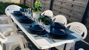 A rectangular outdoor dining table set with white plastic chairs, black plates, and a potted plant.