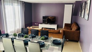 A modern living room with a dining area, purple walls, a flat-screen TV, and a brown sofa.