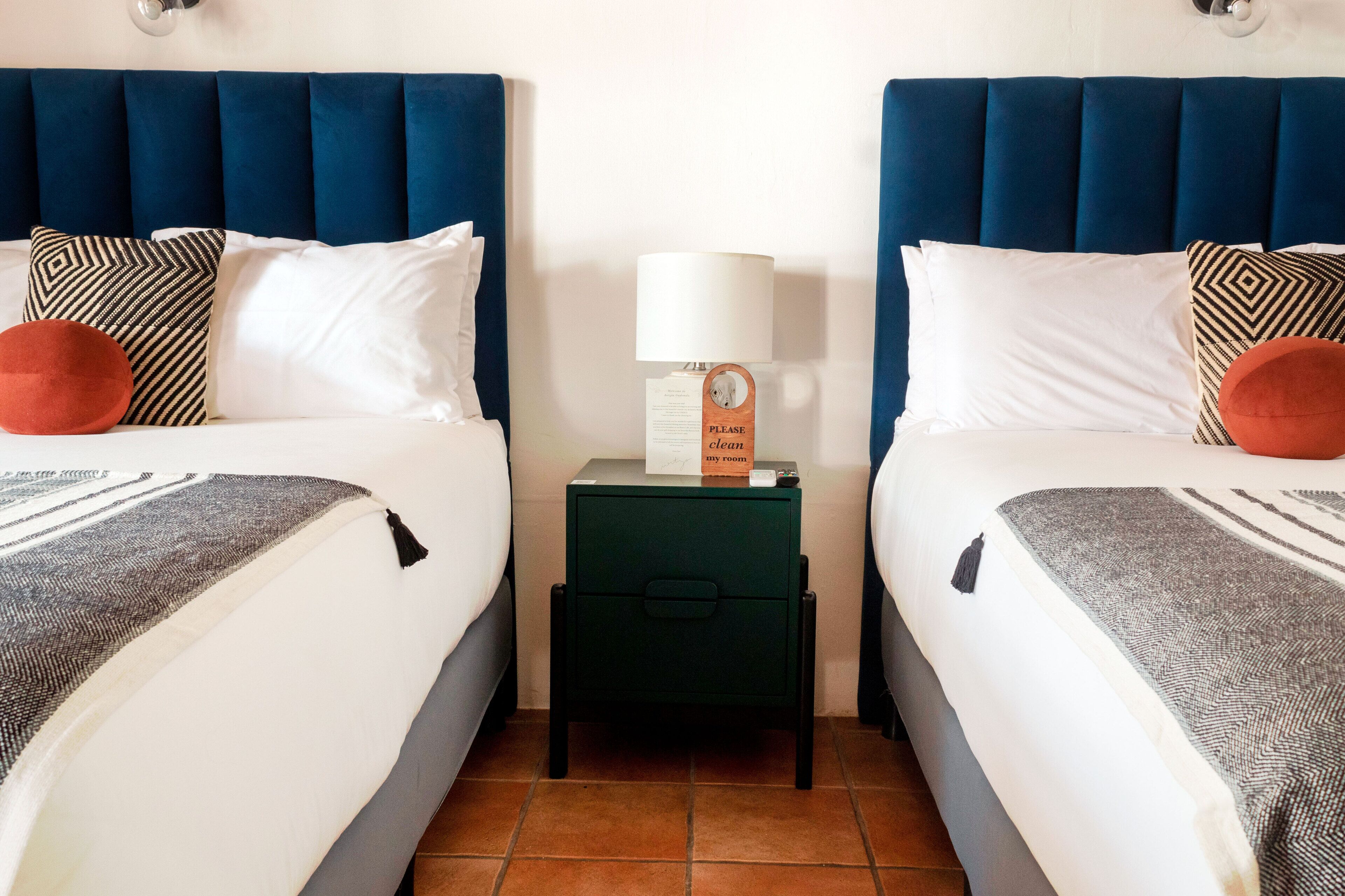 Habitación de hotel con dos camas, cada una con cabecera azul, ropa de cama blanca y almohadas estampadas. Hay una pequeña mesa con una lámpara y un cartel que dice 'Por favor, limpiar' entre las camas.