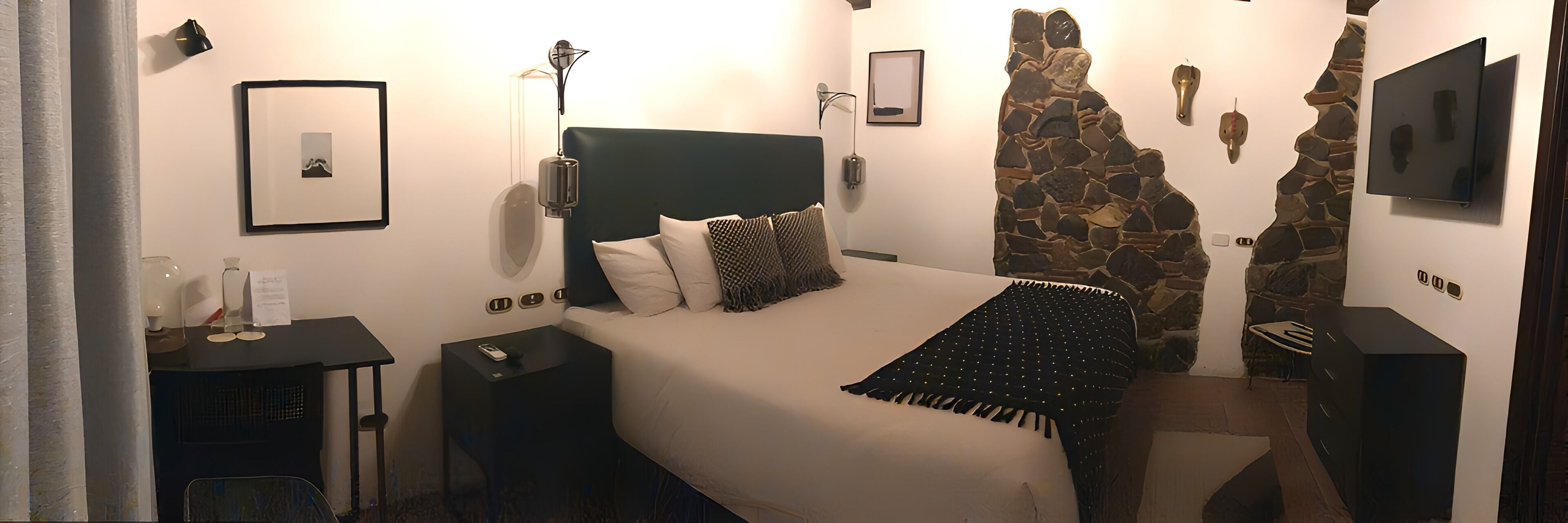 Ein modernes Hotelzimmer mit einem großen Bett, einer Wand aus Stein, einem Flachbildfernseher und einem kleinen Schreibtisch mit Lampe.