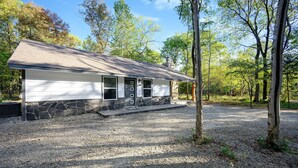 A single-story house with a stone foundation and a sloped roof, surrounded by a gravel driveway and trees. - Romantic honeymoon cabin with hot tub! (Broken Bow)
