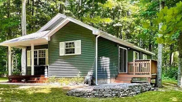 A green single-story house with a porch, stone foundation, and a wooden deck. - 3- Bedroom cottage near Pleasant Lake in Cadillac (Cadillac)