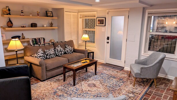 Una sala de estar con un sofá, una mesa de centro, un sillón gris, un tapete estampado y estanterías con diversos objetos.