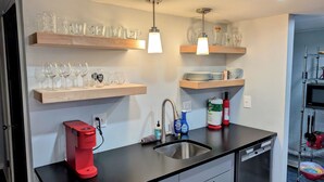 Una cocina con estantes de madera que sostienen cristalería, una cafetera y productos de limpieza.