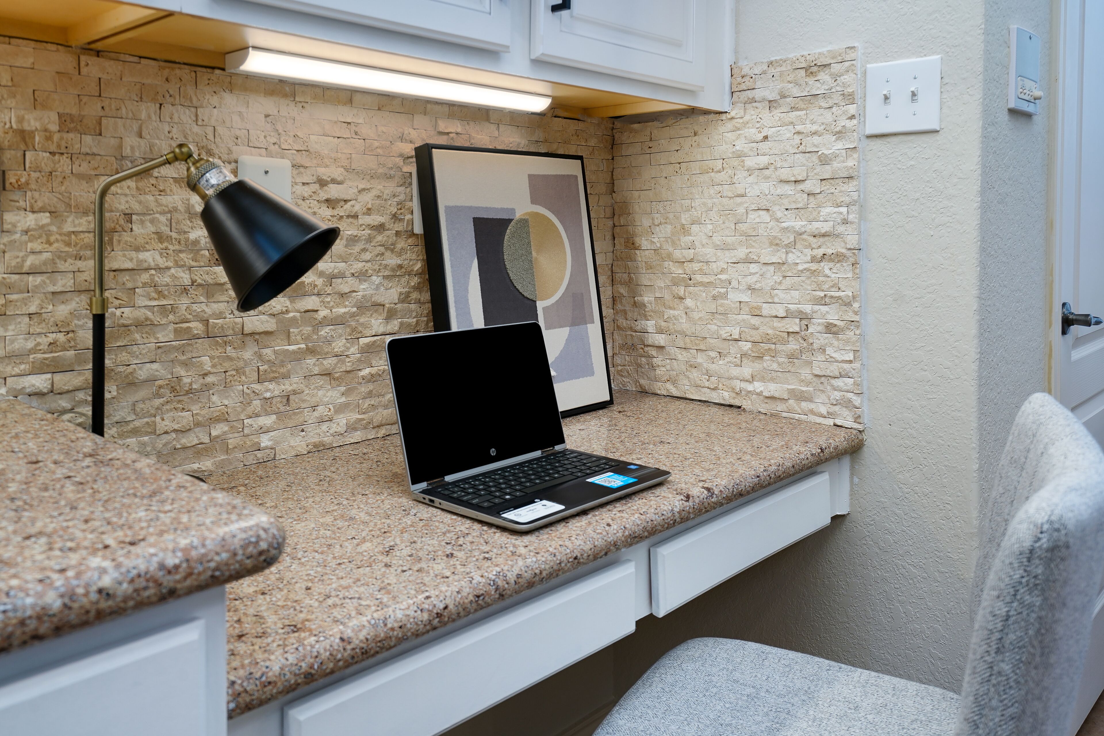 A laptop on a granite countertop, a black desk lamp, and a framed abstract art piece on a brick wall.