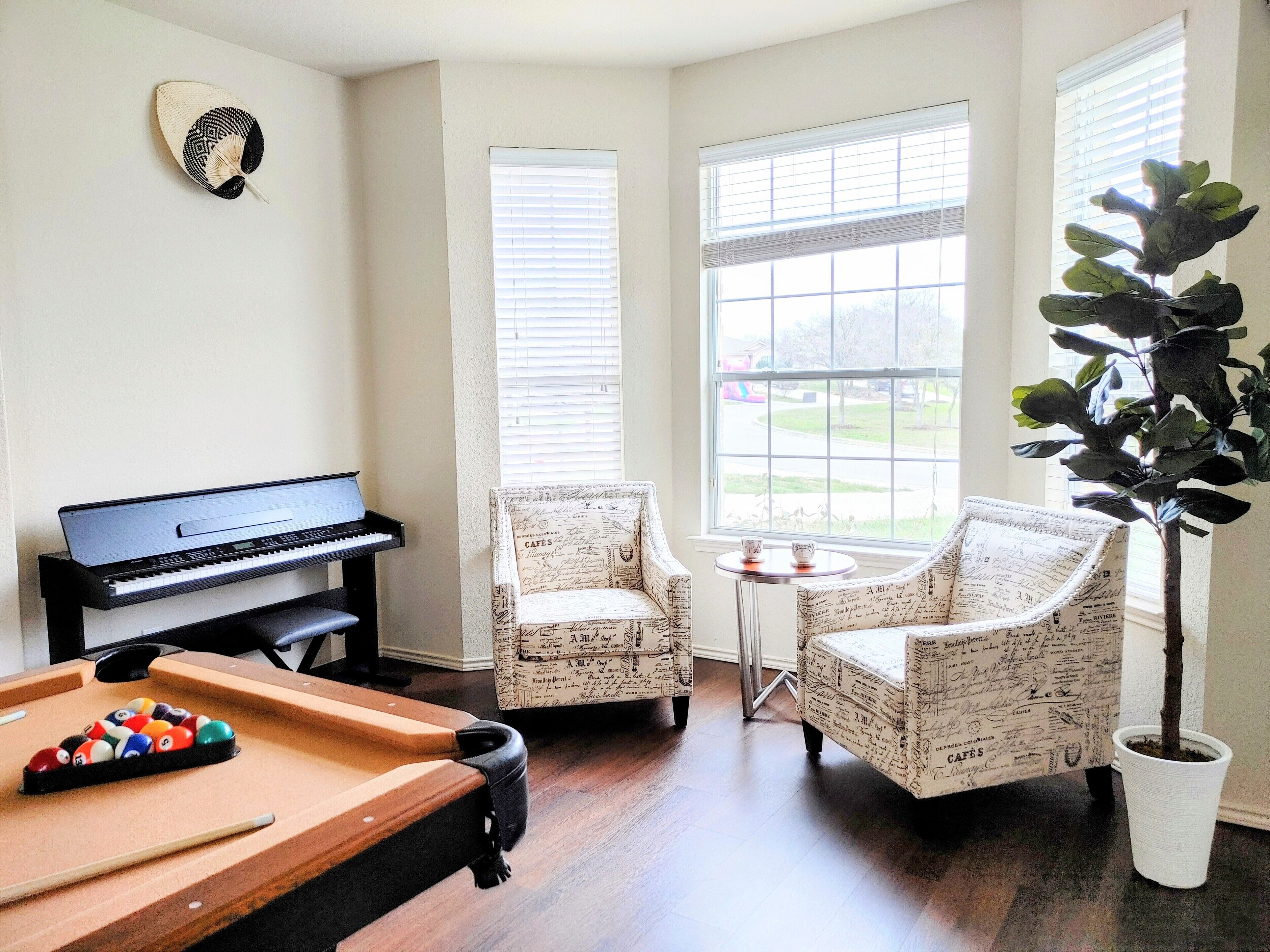 Ein Zimmer mit Poolbillard, einem Klavier, zwei Sesseln, einem kleinen Tisch mit Pflanze und einem Fenster mit Jalousien.