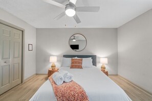 A hotel room with a bed, a ceiling fan, a round mirror, two bedside lamps, and a door.
