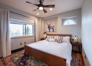 A bedroom with a large bed, a ceiling fan, a window with curtains, a nightstand with a lamp, and a painting on the wall. - Panoramic Views from a Large Peaktop Retreat with Hot Tub, Sauna, & Axe-Throwing (Conifer)