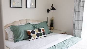 Een net opgemaakt bed met een wit en groen beddegoed, twee kussens en een kussen met stippen. Op het nachtkastje naast het bed staat een zwarte bureaulamp.