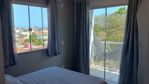 A bedroom with a bed, large windows, and a view of buildings and trees. - Suite with beautiful sea view!! (Florianópolis)