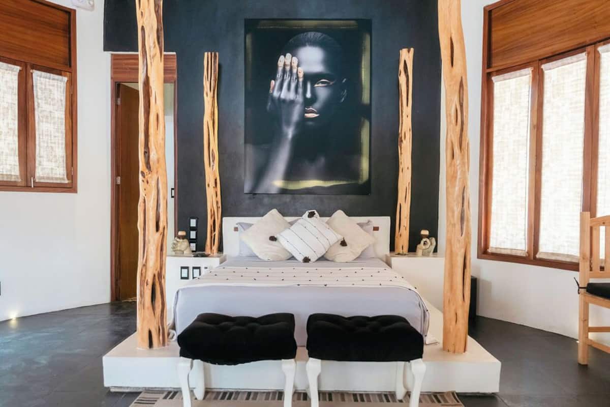 A modern bedroom with a large bed, two black ottomans, a painting above the bed, and wooden elements.