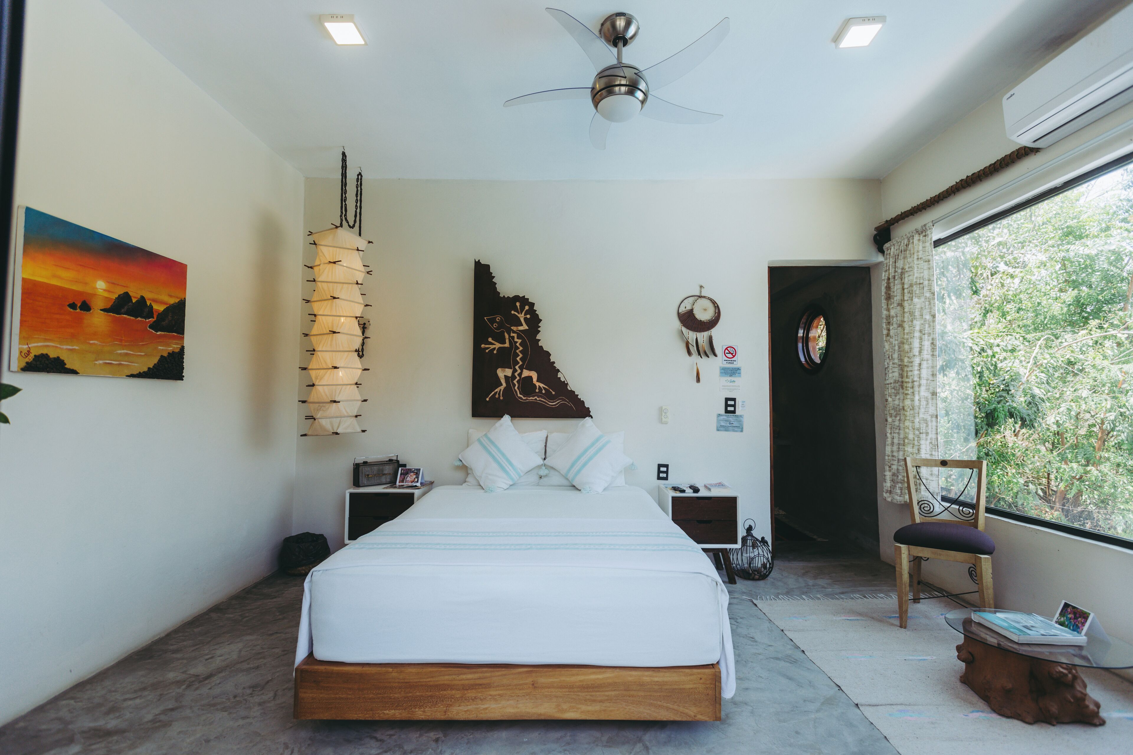 A bedroom with a bed, a ceiling fan, a nightstand, a chair, and a window with a view of greenery.