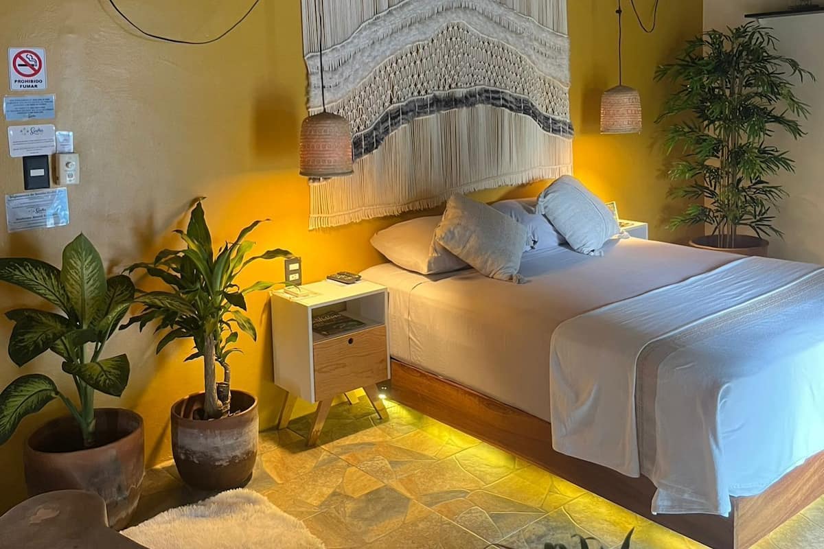 A bedroom with a bed, nightstands, a hanging macrame wall hanging, a potted plant, and a thatched roof ceiling.