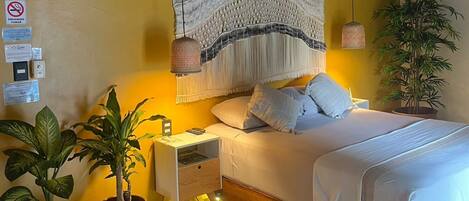 A bedroom with a bed, nightstands, a hanging macrame wall hanging, a potted plant, and a thatched roof ceiling.