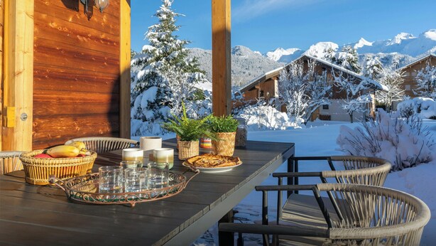 A wooden deck with a table set for dining, featuring a basket of fruit, candles, and a tray. - Chalet Antigone - ski holiday for 8 with mountain view - OVO Network (Saint-Jean-de-Sixt)
