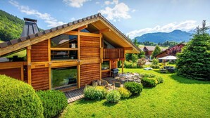 A wooden cabin with a deck, surrounded by lush greenery and mountains in the background. - Chalet Antigone - ski holiday for 8 with mountain view - OVO Network (Saint-Jean-de-Sixt)
