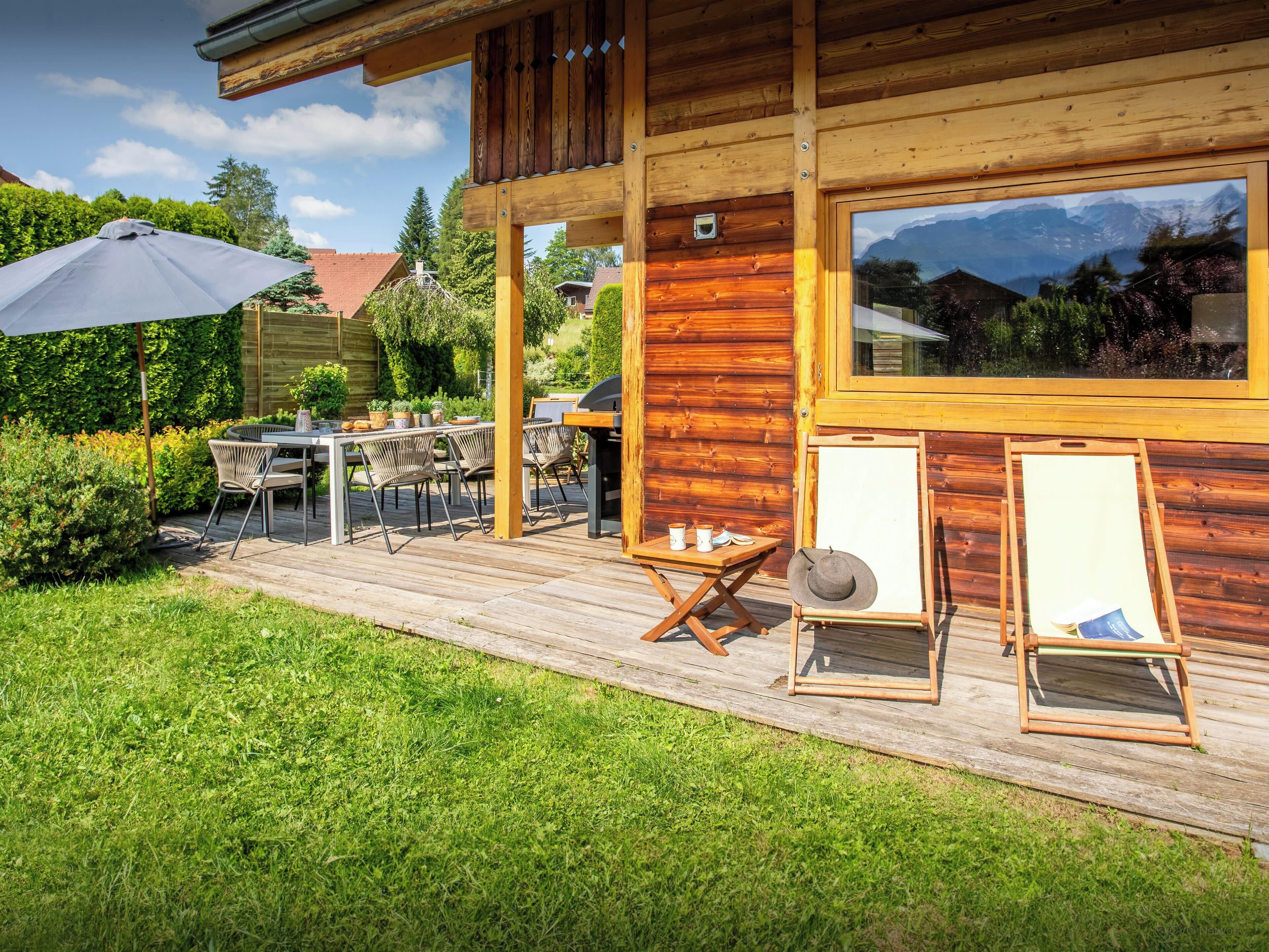 A wooden deck with outdoor furniture, a grill, and a large window overlooking a mountainous landscape.