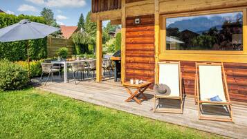 A wooden deck with outdoor furniture, a grill, and a large window overlooking a mountainous landscape.