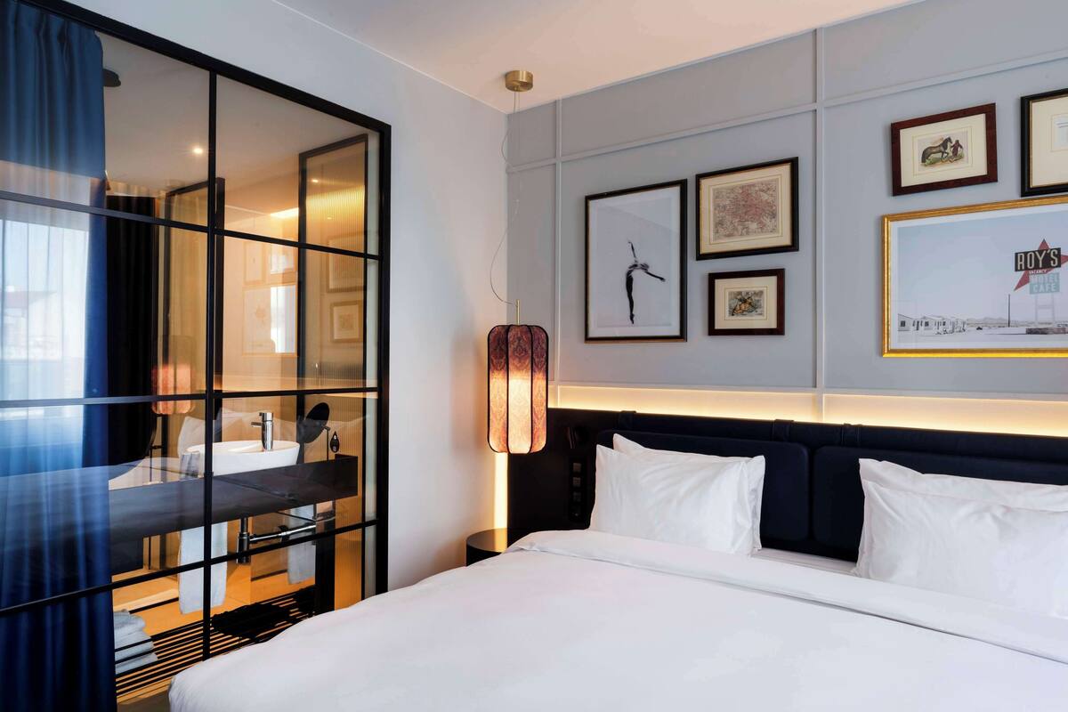 A hotel room with a large bed, a bedside table, a mirror, a wall-mounted lamp, and framed pictures on the wall.