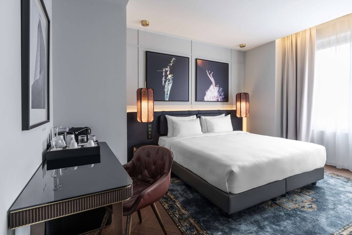 A hotel room with a bed, a desk with a coffee machine, two framed pictures on the wall, and a chair.