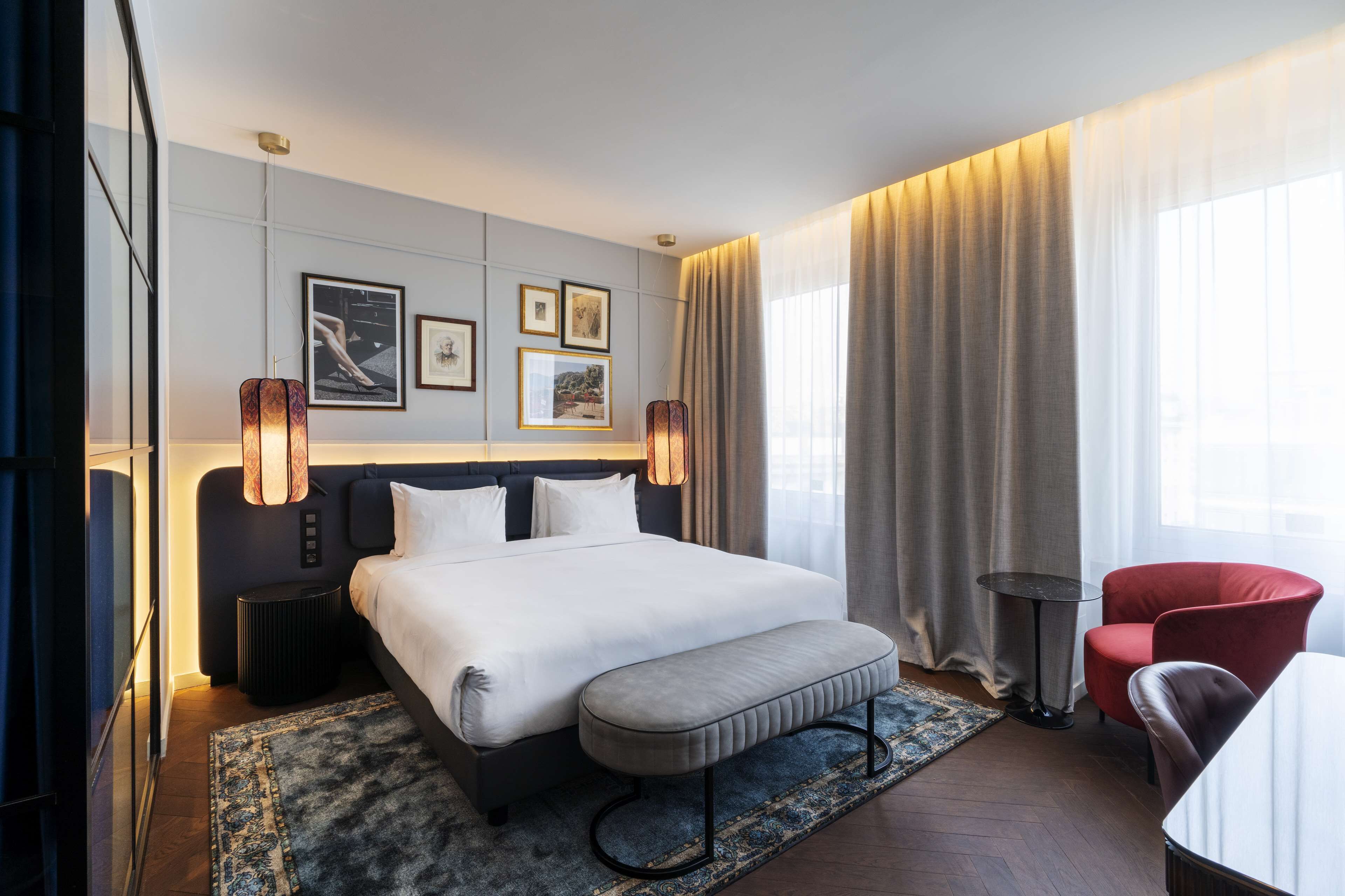 Een moderne hotelkamer met een groot bed, een bankje, een rode fauteuil, een kleine tafel en een groot raam met gordijnen.
