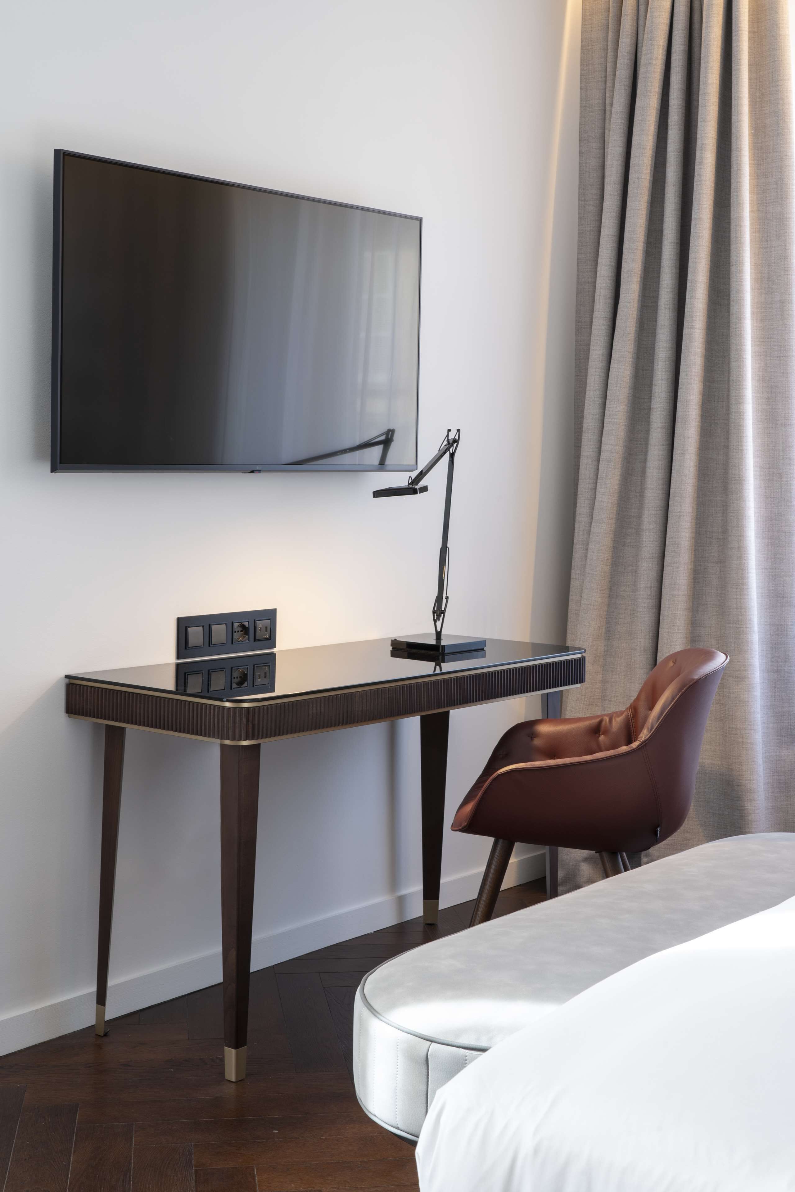 A modern hotel room with a flat-screen TV mounted on the wall, a desk with a lamp, and a leather chair.