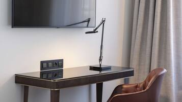 A modern hotel room with a flat-screen TV mounted on the wall, a desk with a lamp, and a leather chair.